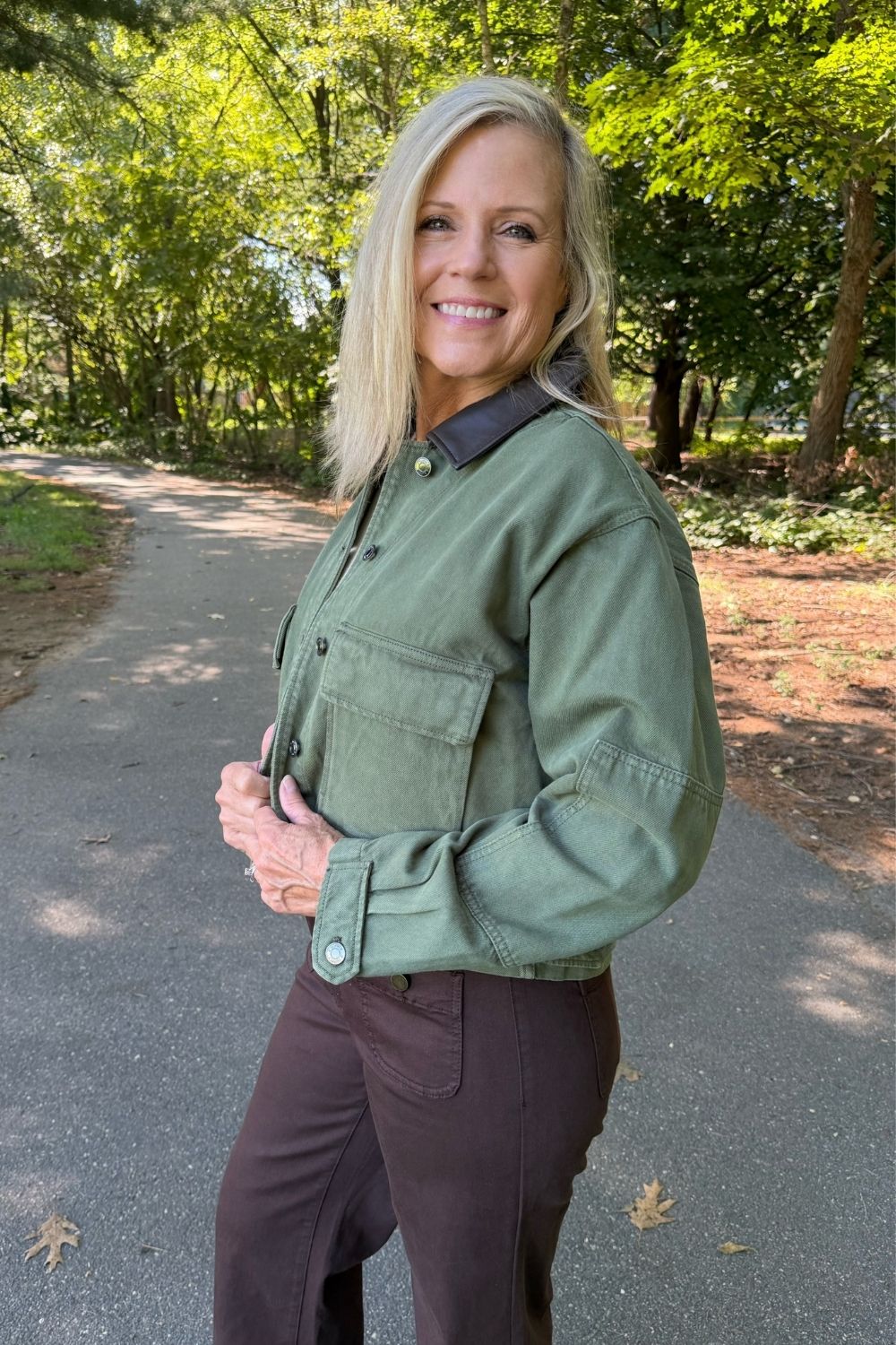 A smiling blonde woman stands on a paved path outdoors wearing an olive green utility-style button-up shirt with a dark collar and dark brown pants.