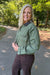 A smiling blonde woman stands on a paved path outdoors wearing an olive green utility-style button-up shirt with a dark collar and dark brown pants.