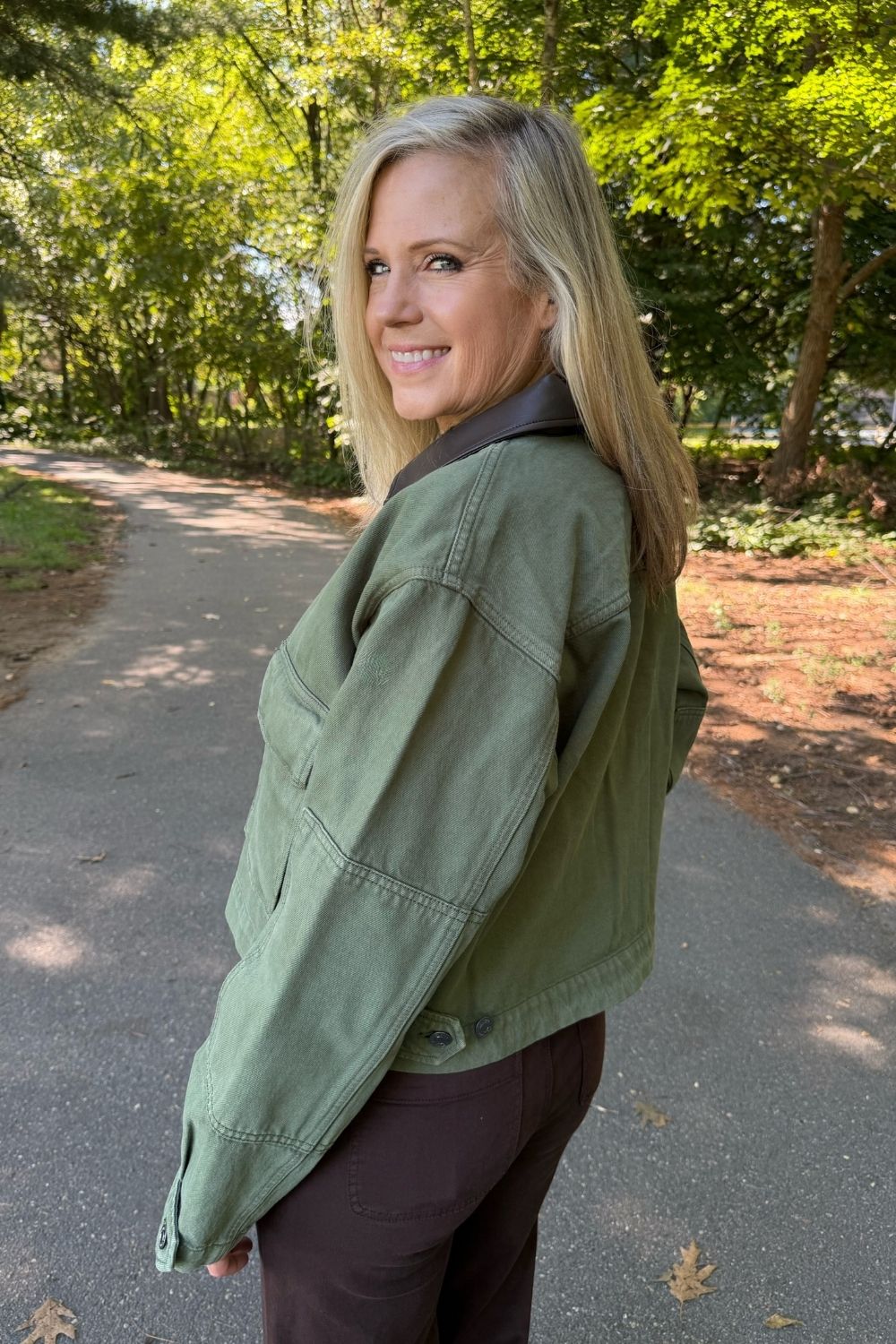 A woman with blonde hair stands on a paved path outdoors, wearing an olive green cropped jacket with dark brown pants. Trees and foliage are visible in the background.