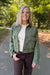 A woman standing on a paved path in a wooded outdoor setting, wearing an olive green cropped utility jacket over a cream ribbed sweater and dark brown pants.