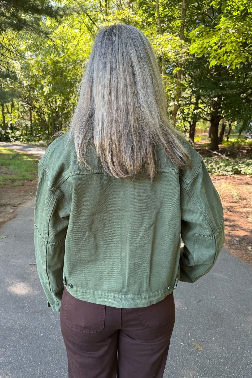 A person viewed from behind wearing an olive green canvas utility jacket and brown pants, standing on a paved path in a wooded outdoor setting.