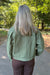 A person viewed from behind wearing an olive green canvas utility jacket and brown pants, standing on a paved path in a wooded outdoor setting.