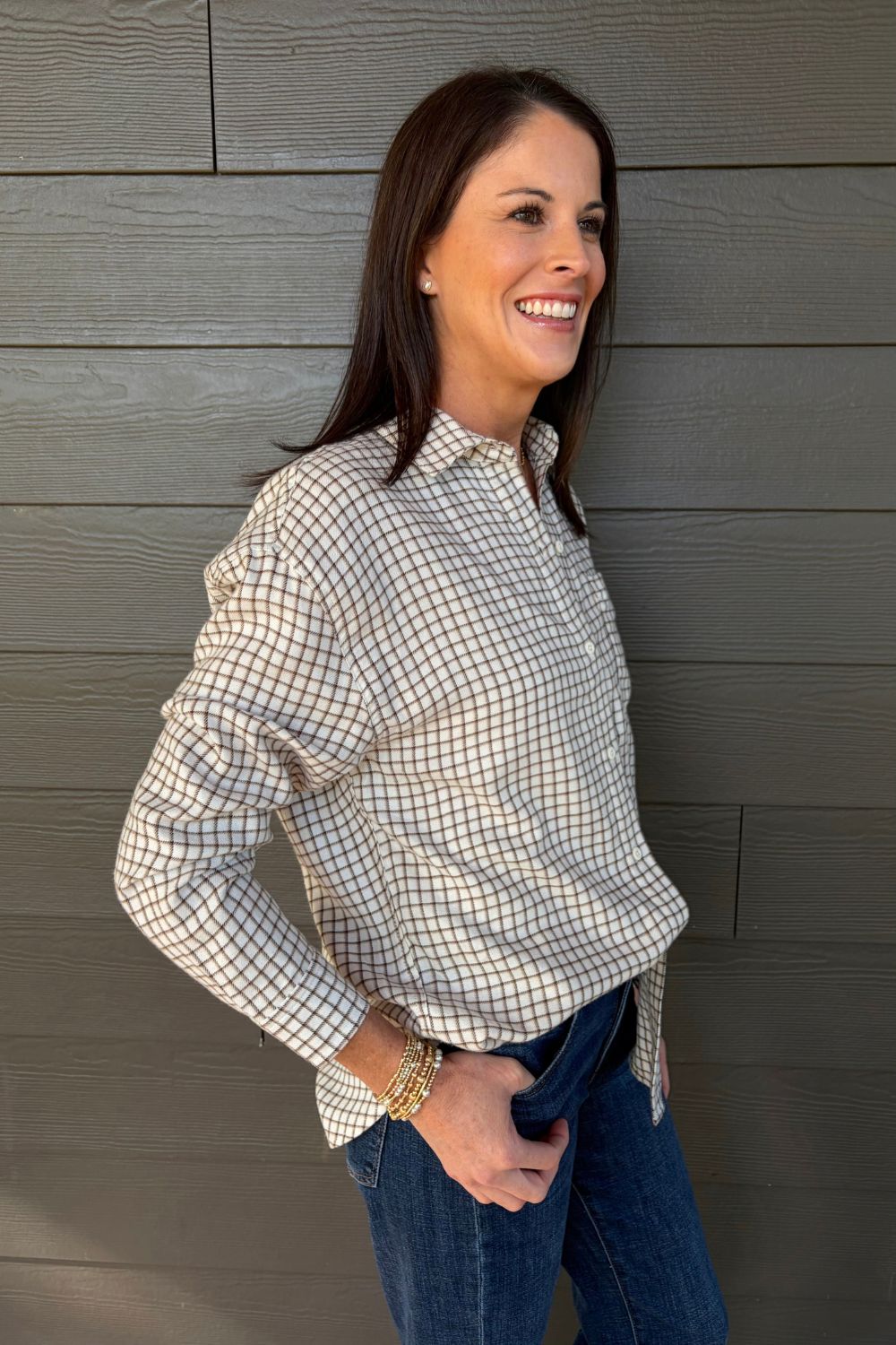 A woman smiling and looking to the side, wearing a beige and white checkered long-sleeve button-down shirt and blue jeans, standing against a gray wood-paneled wall.