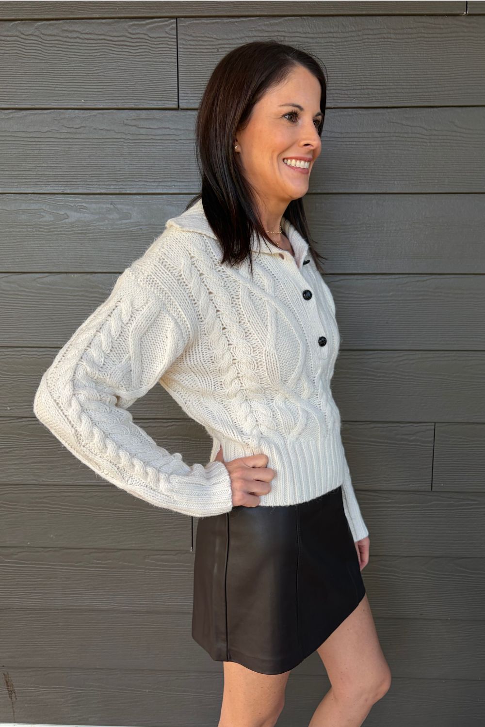 A woman shown from the side wearing a cream cable-knit sweater and a dark brown mini skirt, standing against a gray wood-paneled wall.