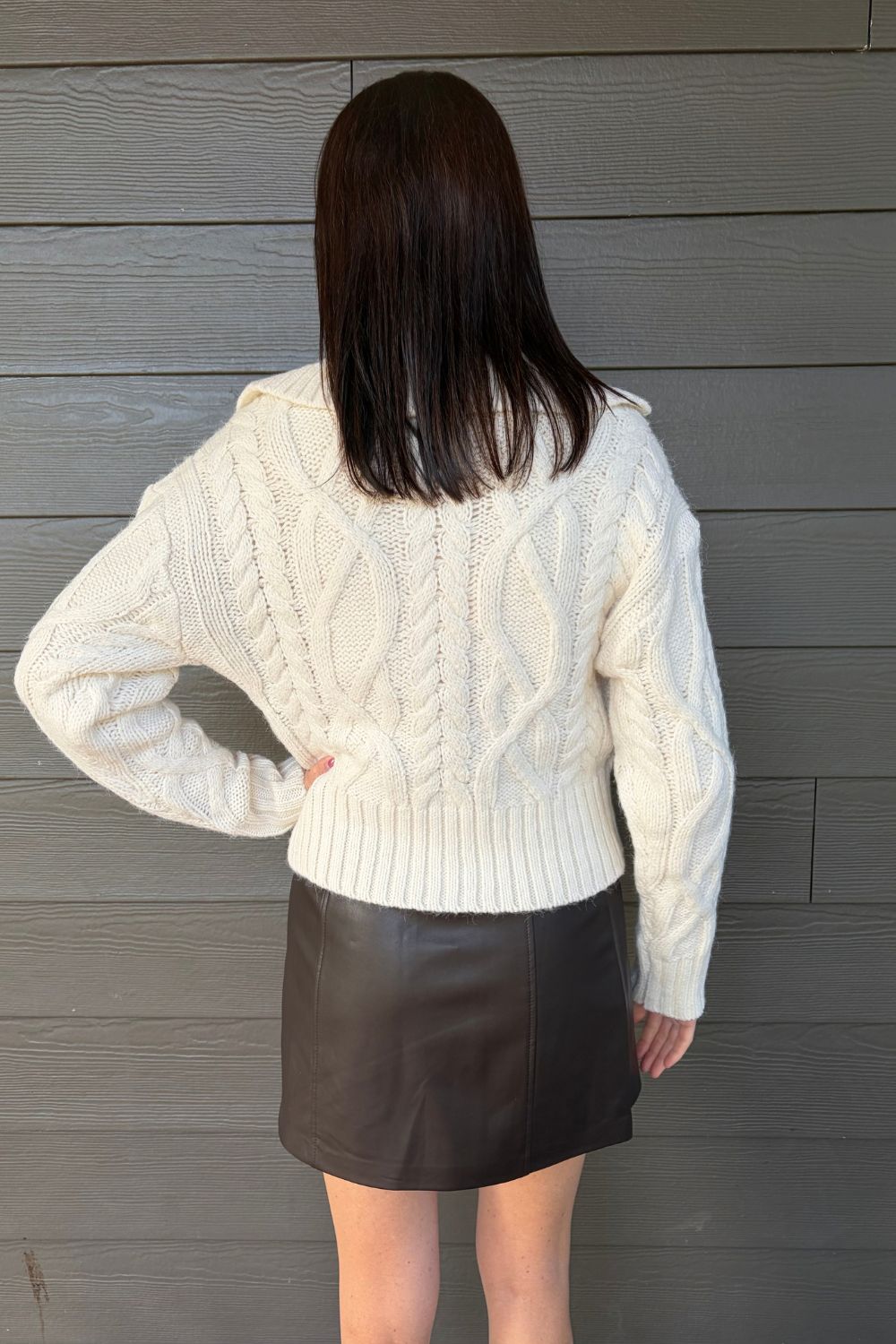 A woman shown from behind wearing a cream cable-knit sweater and a dark brown mini skirt, standing against a gray wood-paneled wall.