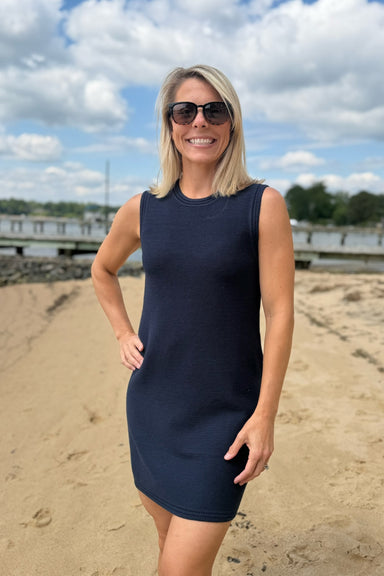 Woman in a navy dress standing on a sandy beach with a blue sky and clouds in the background.