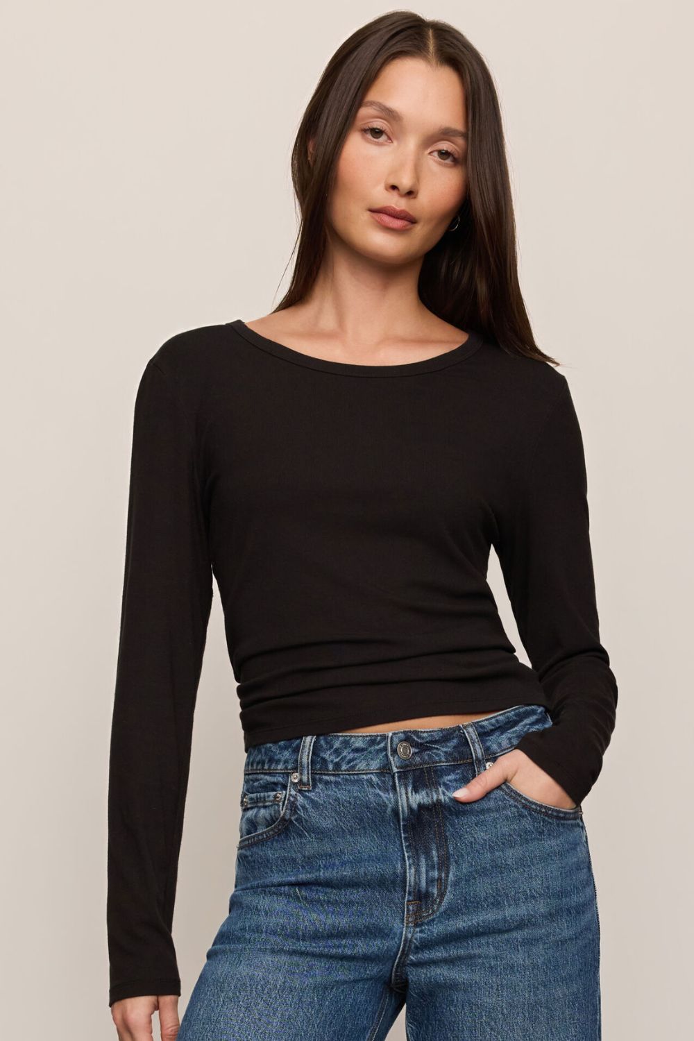 A woman modeling a black long-sleeve crewneck top paired with blue jeans, photographed against a plain light beige studio background.