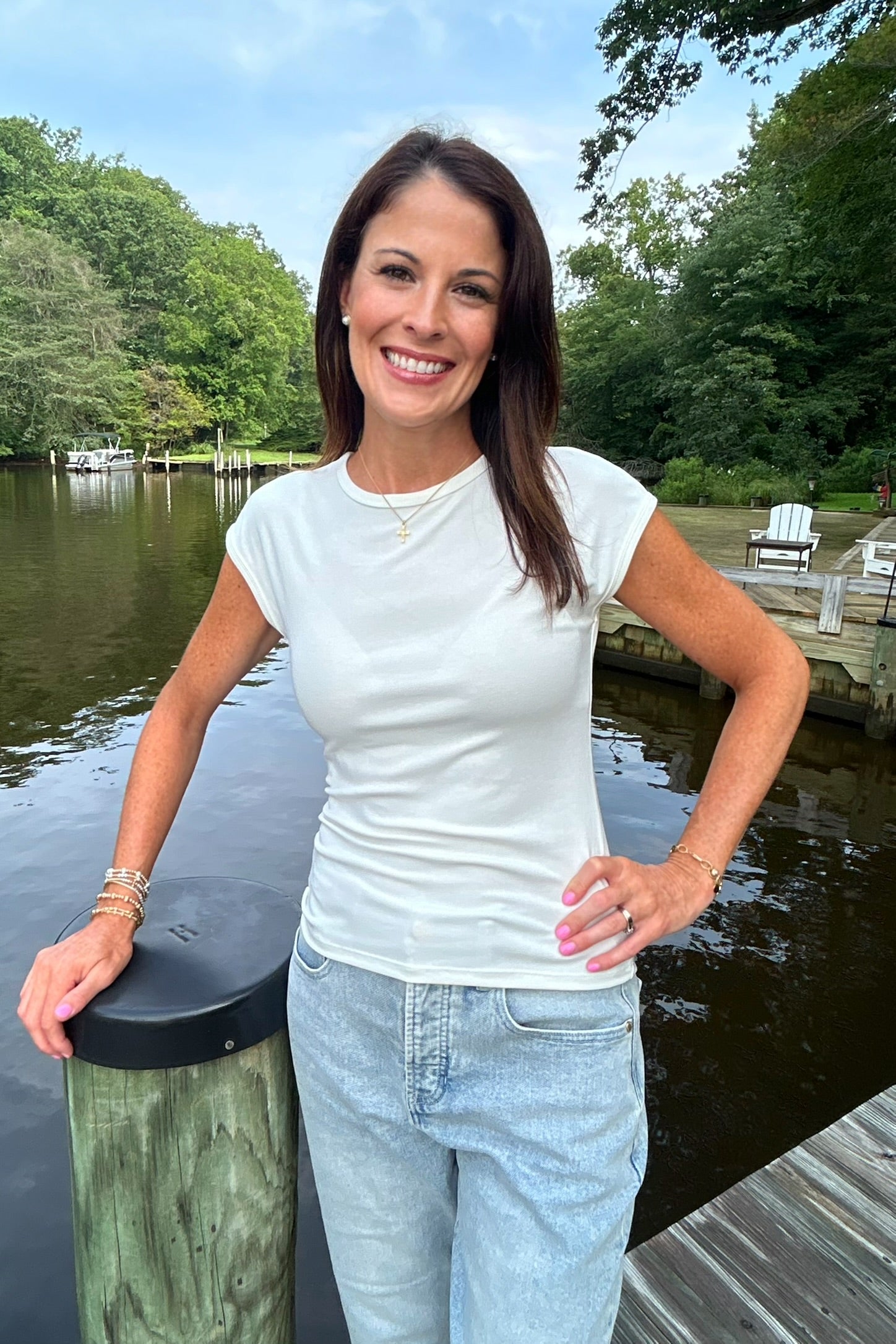 A woman smiling and posing outdoors by a lake dock, wearing a fitted white short-sleeve crewneck t-shirt and light wash blue jeans.