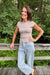 A woman posing outdoors on a wooden boardwalk wearing a beige short-sleeve crewneck top and light-wash blue jeans, with a lush green forest background.
