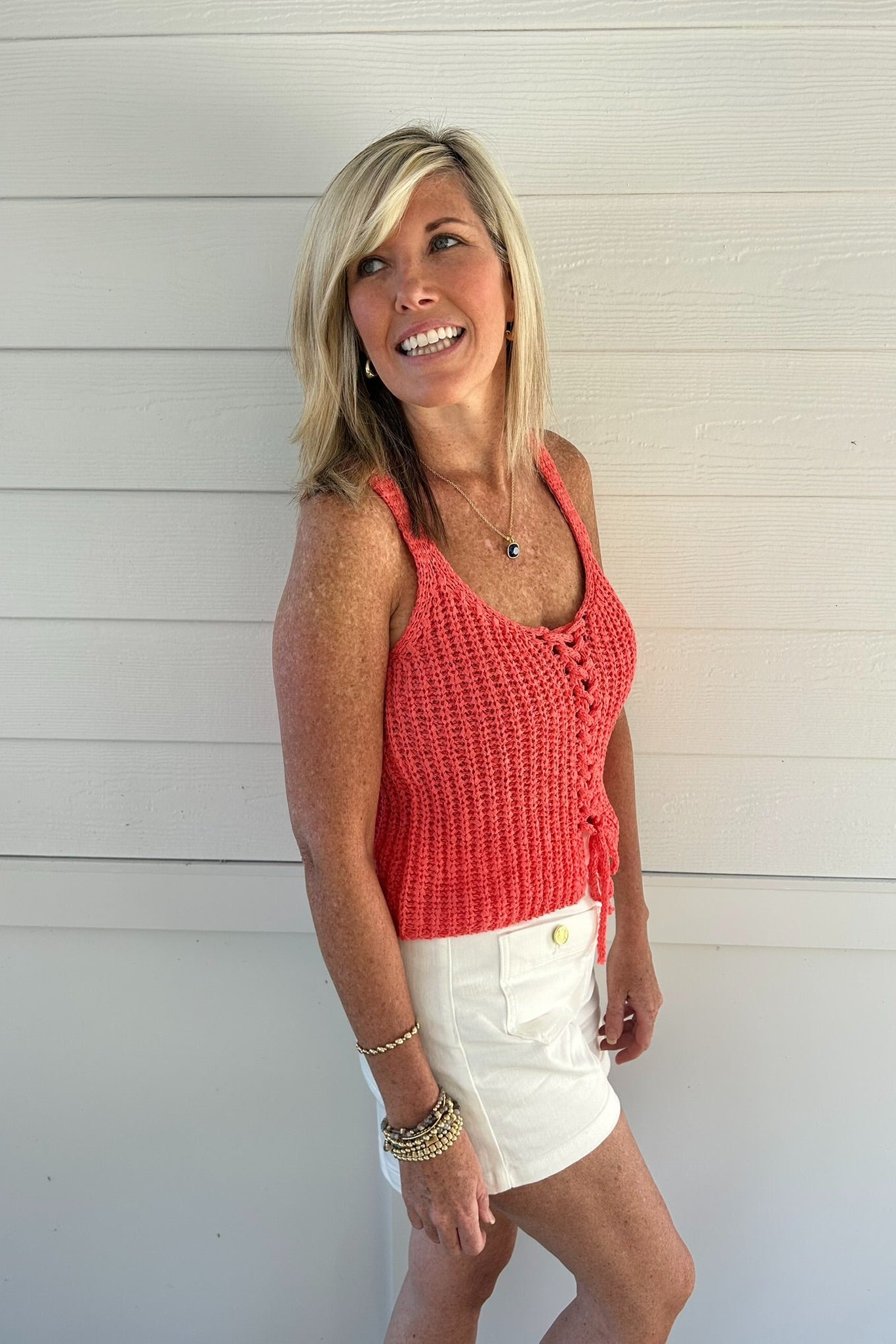 Woman wearing a pink crochet tank top and white shorts standing against a light wooden wall.