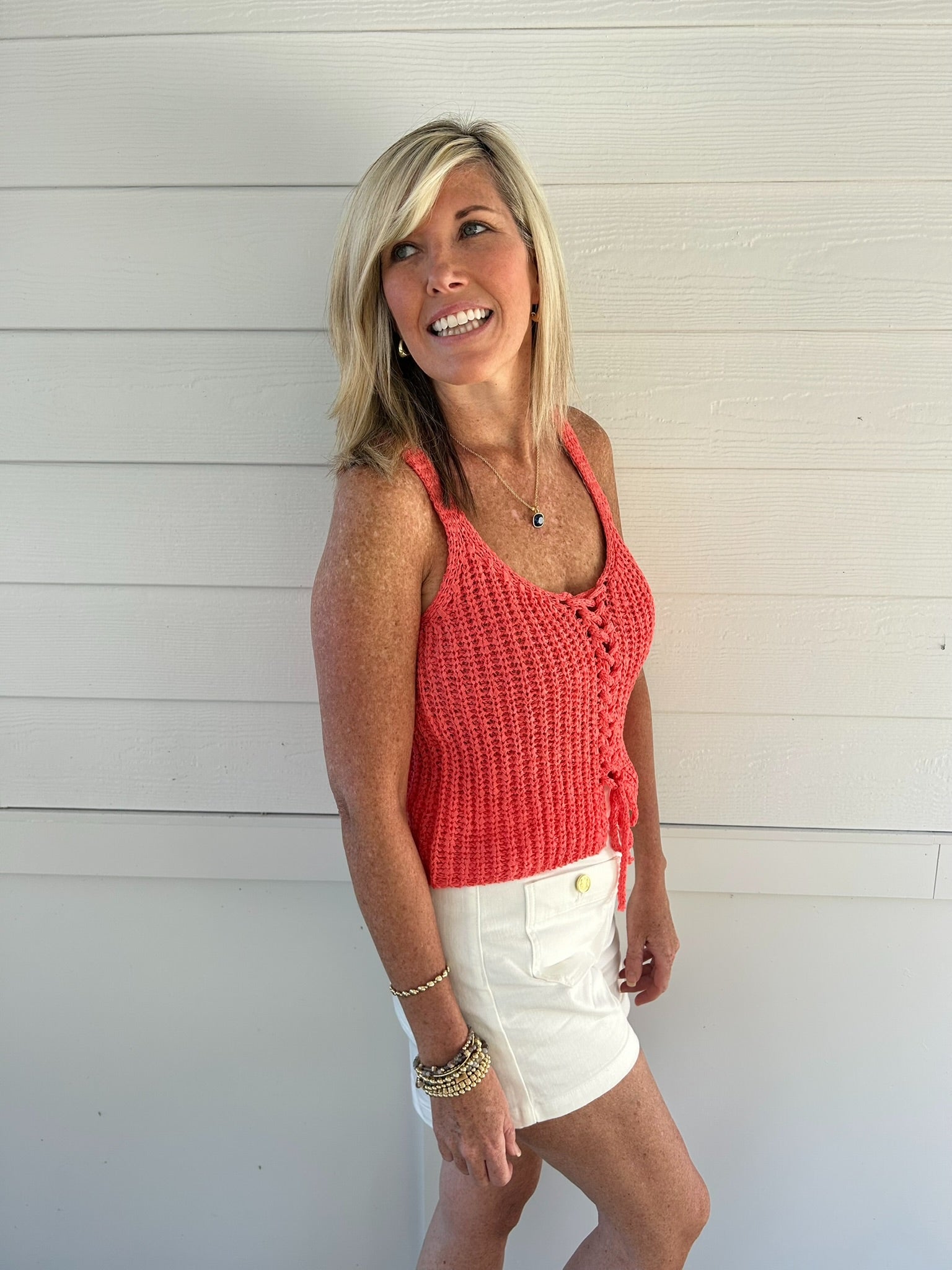 Woman wearing a pink crochet tank top and white shorts standing against a light wooden wall.