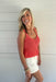 Woman wearing a pink crochet tank top and white shorts standing against a light wooden wall.