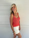 Woman wearing a pink crochet tank top and white shorts standing against a light wooden wall.