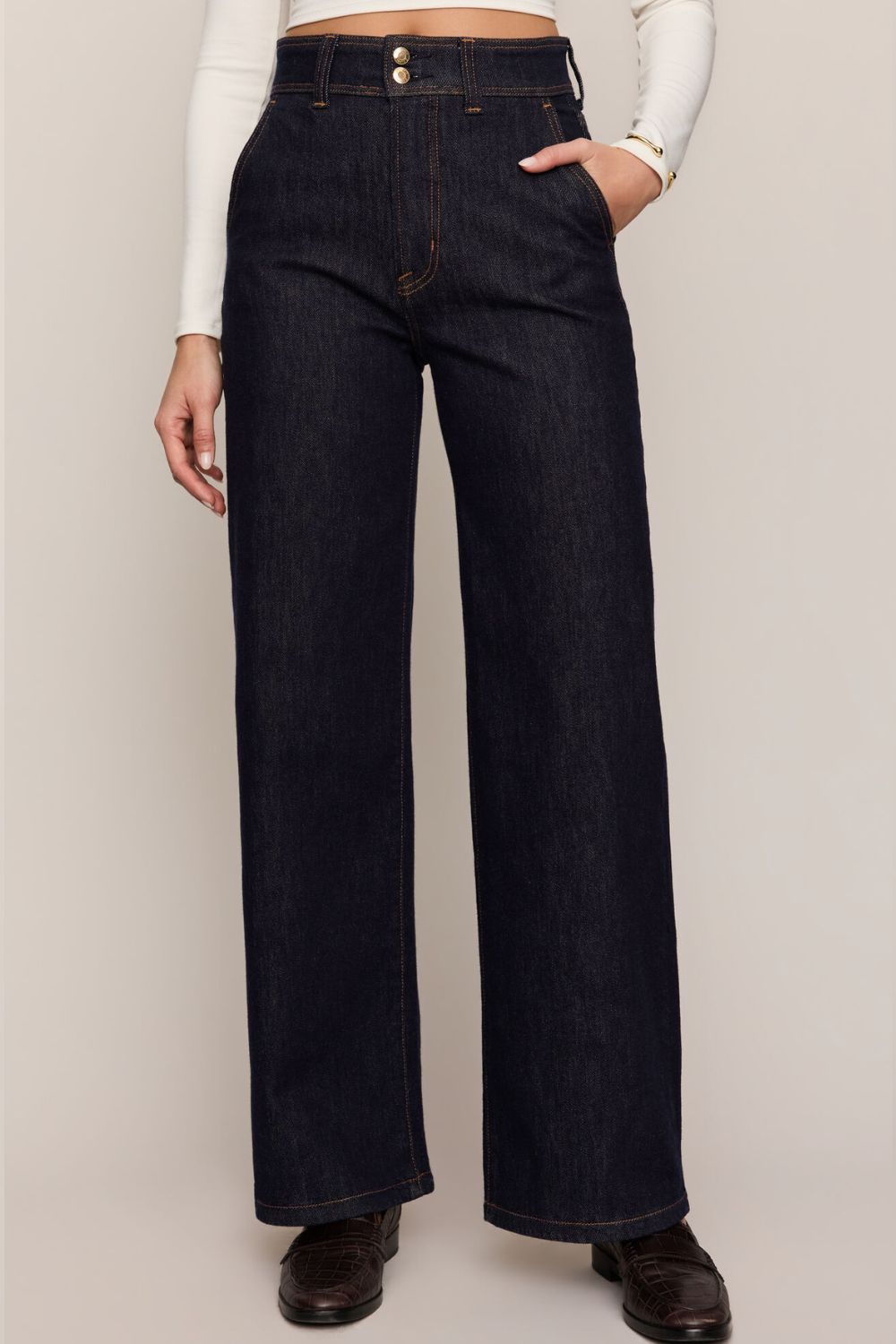 A person wearing dark indigo wide-leg denim jeans with a white long-sleeve top and dark brown leather shoes, shown from the waist down against a plain light beige studio background.