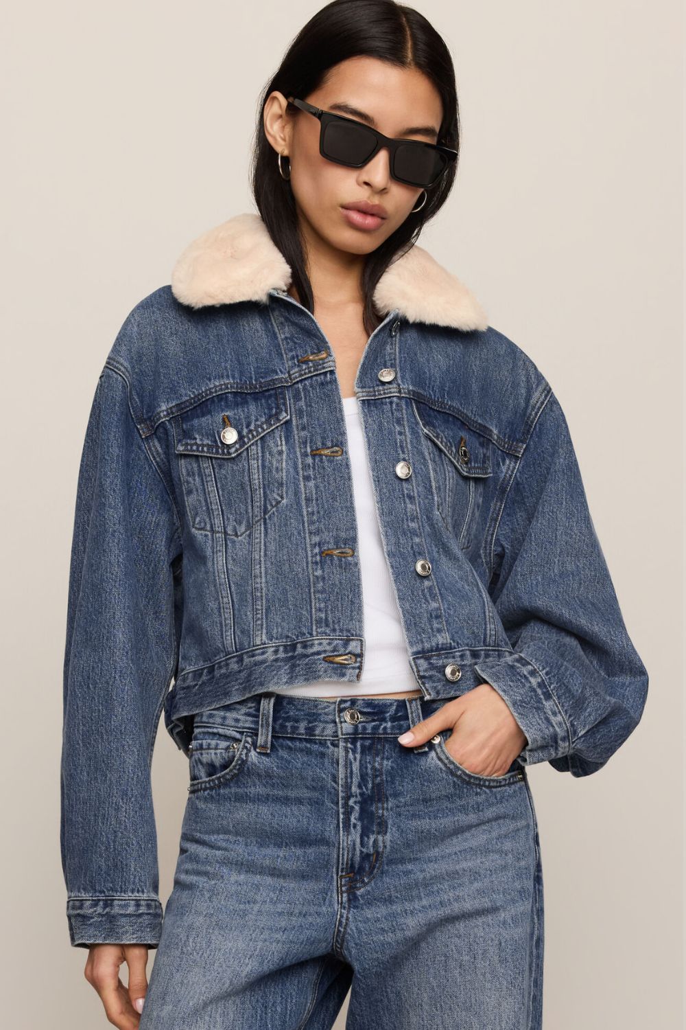 A model wearing a cropped blue denim jacket with a cream shearling-style collar, paired with matching blue jeans and black sunglasses, against a solid light beige background.