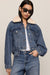 A model wearing a cropped blue denim jacket with a cream shearling-style collar, paired with matching blue jeans and black sunglasses, against a solid light beige background.