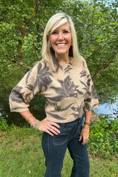 A smiling blonde woman wearing a beige knit top with a dark brown floral pattern and dark blue jeans, standing outdoors in front of a green wooded background.