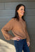 A woman wearing a brown long-sleeve henley sweater and blue jeans, standing against a gray wood-paneled wall.