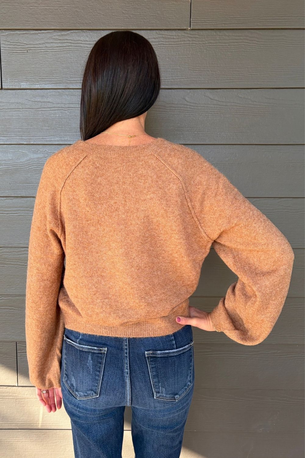 A woman viewed from behind wearing a fuzzy, heathered camel-colored crewneck sweater and blue jeans, standing against a gray wood-paneled wall.