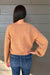 A woman viewed from behind wearing a fuzzy, heathered camel-colored crewneck sweater and blue jeans, standing against a gray wood-paneled wall.