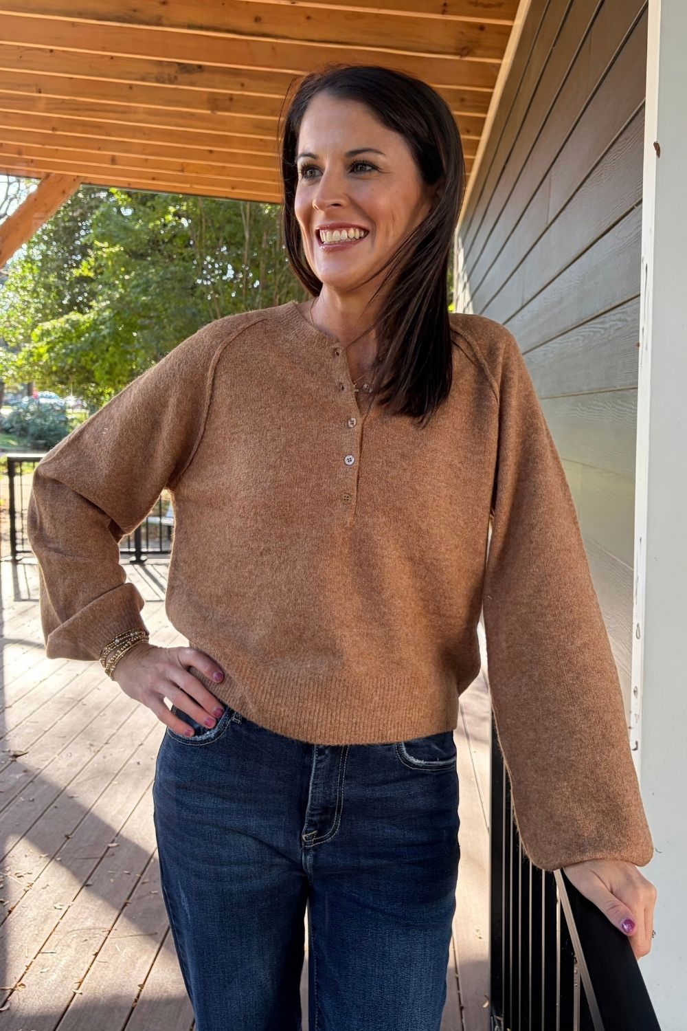 A woman wearing a warm brown long-sleeve henley knit sweater and dark blue jeans, standing on a wooden porch.