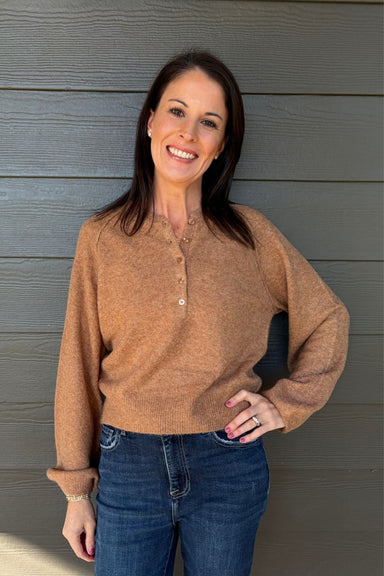 A woman smiling and posing in front of a gray wood-paneled wall, wearing a fuzzy tan-brown long-sleeve henley sweater and blue jeans.