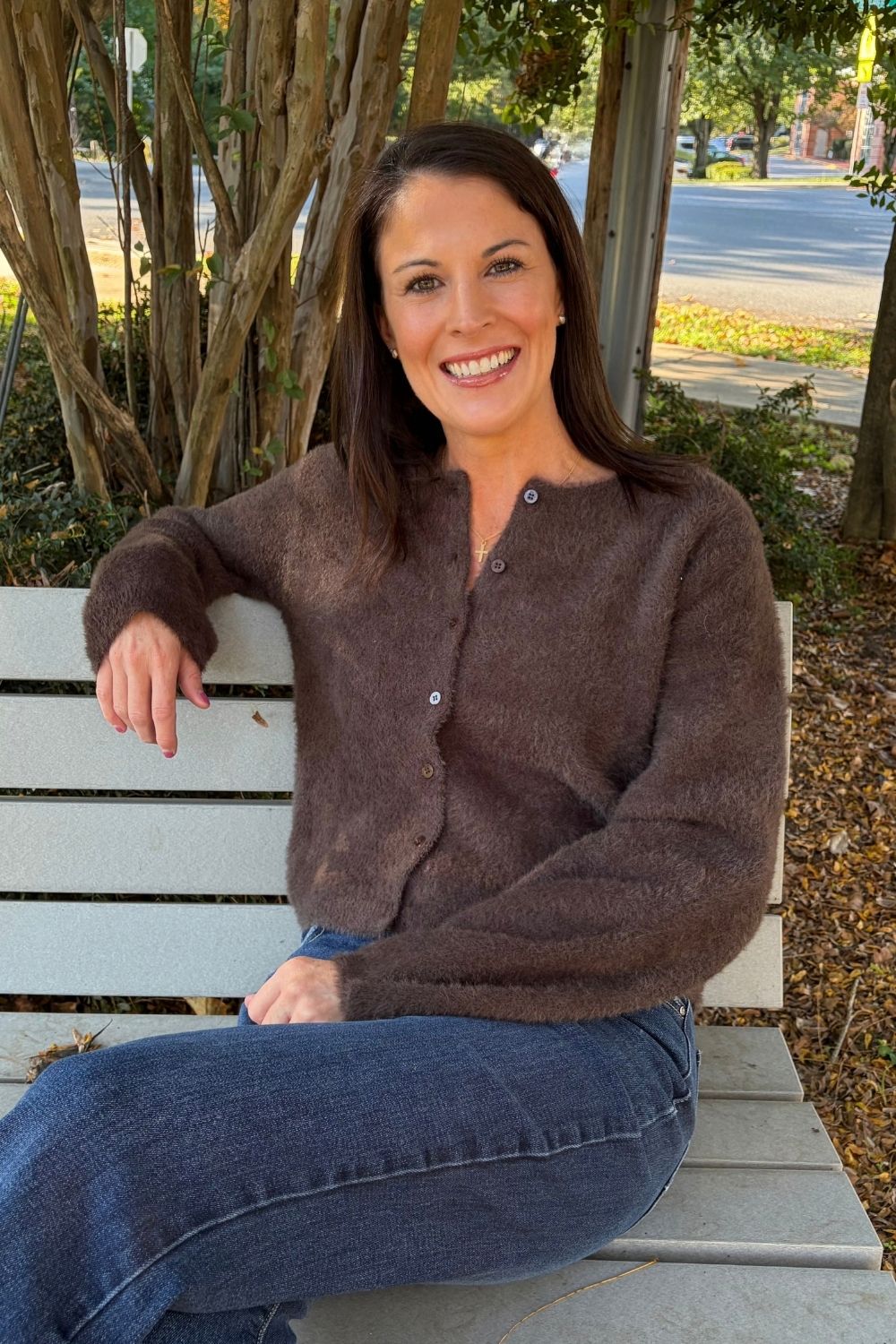 A woman smiling while sitting on a white wooden bench outdoors. She is wearing a fuzzy, long-sleeve brown button-front cardigan and blue jeans, with trees and a street visible in the background.