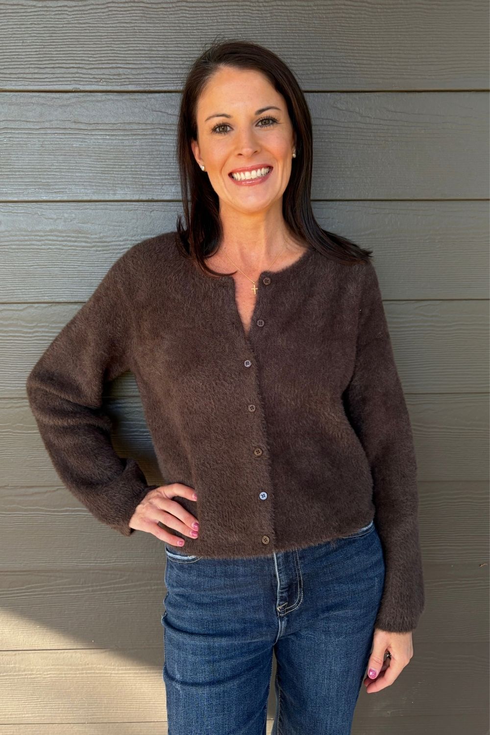 A smiling woman wearing a brown fuzzy long-sleeve button-front cardigan and blue jeans, standing against a gray wood-paneled wall.