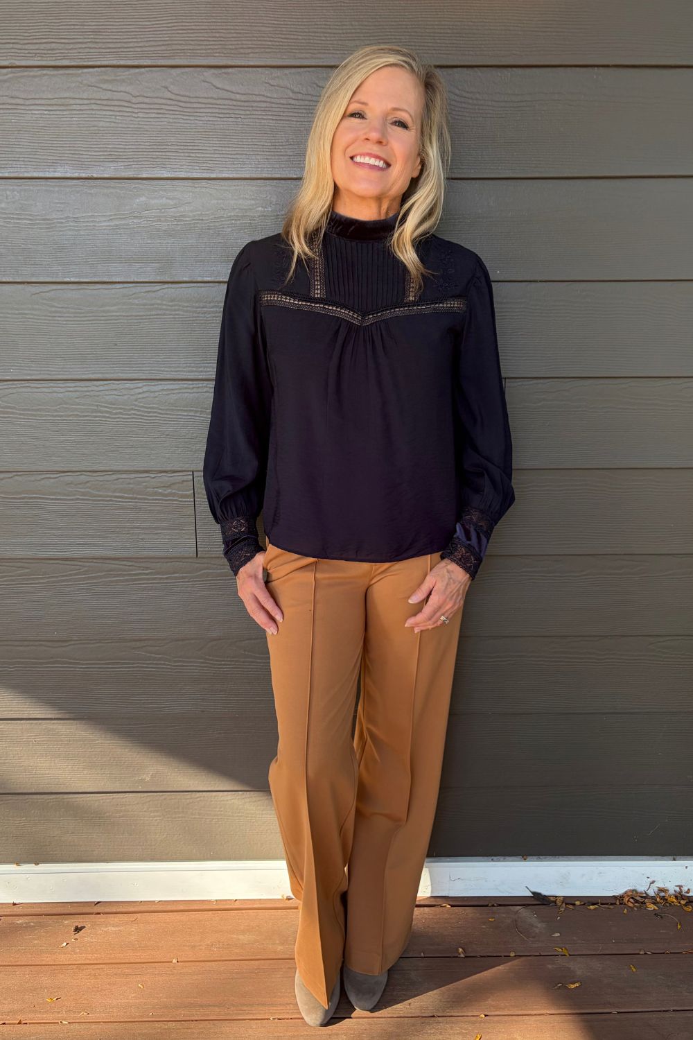 A woman stands on a wooden deck in front of a gray wood-paneled wall, wearing a dark navy mock-neck blouse with textured yoke detailing and wide-leg tan pants.