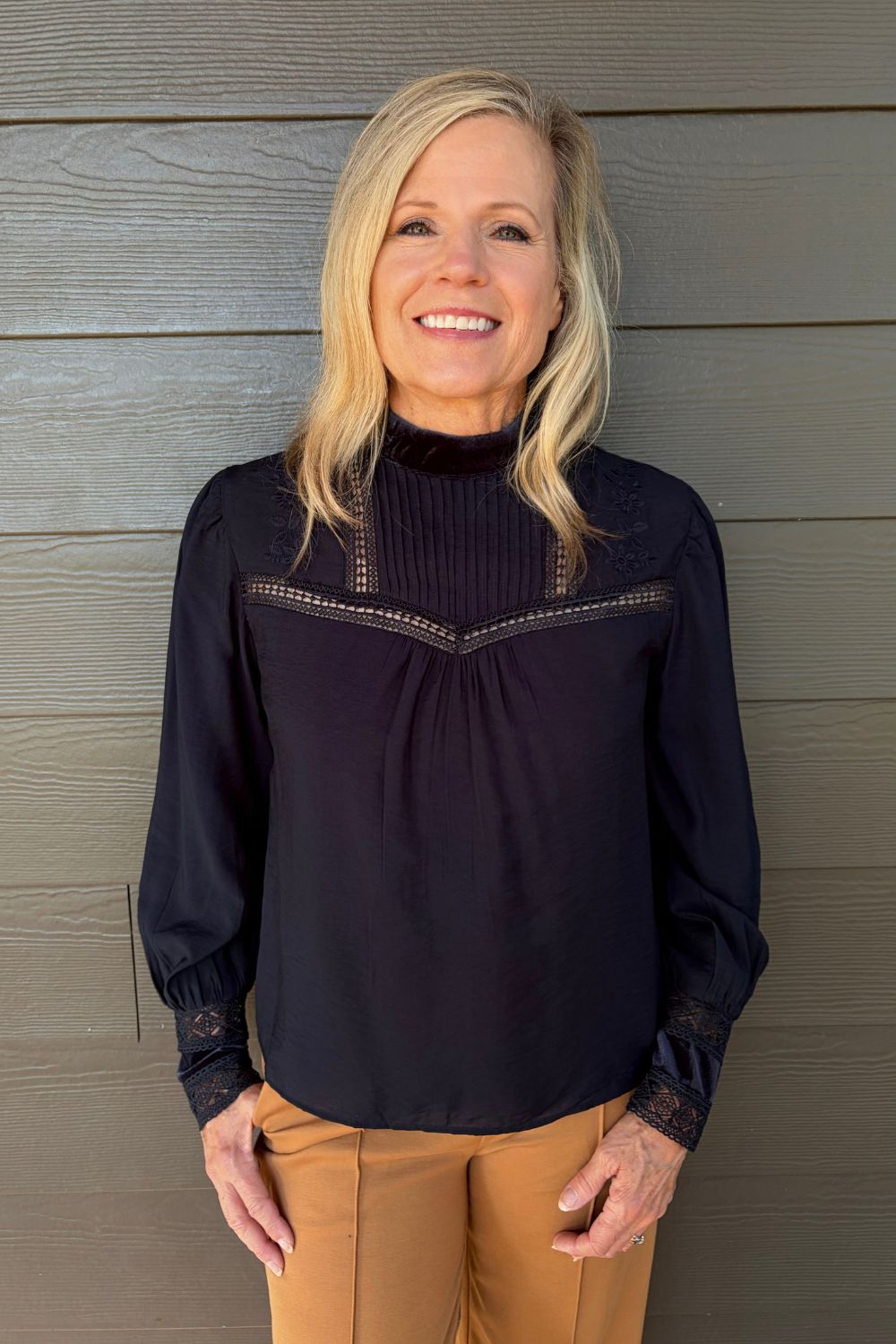 A smiling blonde woman wearing a dark navy long-sleeve blouse with lace trim and tan pants, standing against a gray wood-paneled wall.