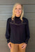 A smiling blonde woman wearing a dark navy long-sleeve blouse with lace trim and tan pants, standing against a gray wood-paneled wall.