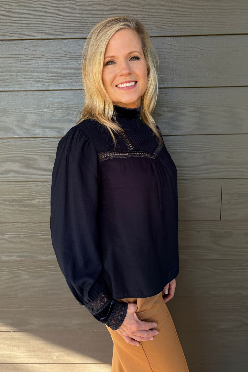 A smiling blonde woman wearing a dark navy long-sleeve blouse with lace detailing and tan pants, standing against a gray wood-paneled wall.
