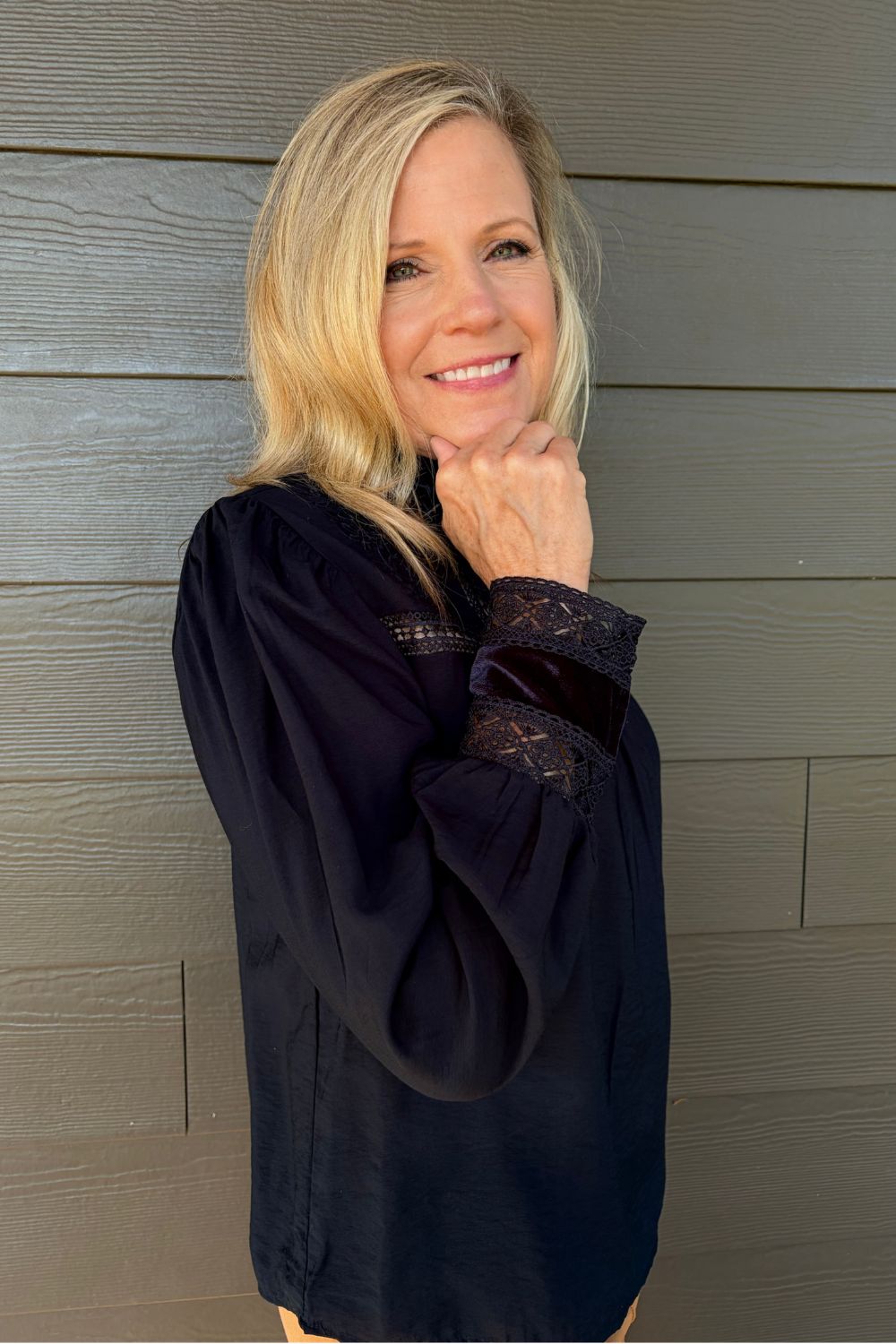 A smiling blonde woman wearing a dark navy long-sleeve blouse with lace cuffs, posing against a gray wood-paneled wall.