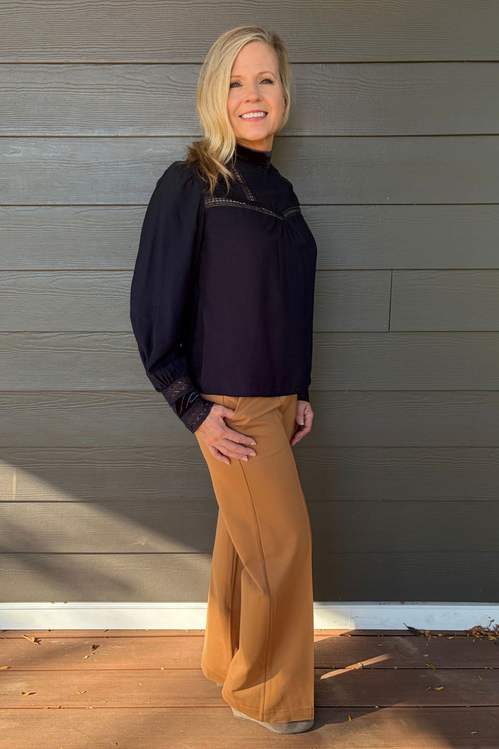 A smiling blonde woman wearing a dark navy long-sleeve blouse with lace detailing and tan pants, standing against a gray wood-paneled wall.