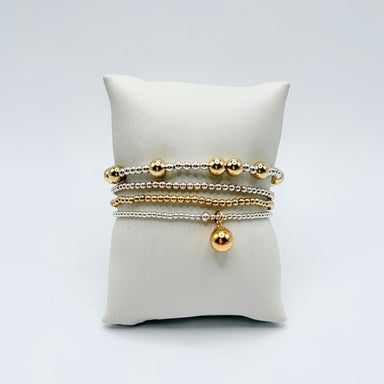 Four mixed metal bracelets in silver and gold beads on a white pillow with a dangling gold ball charm
