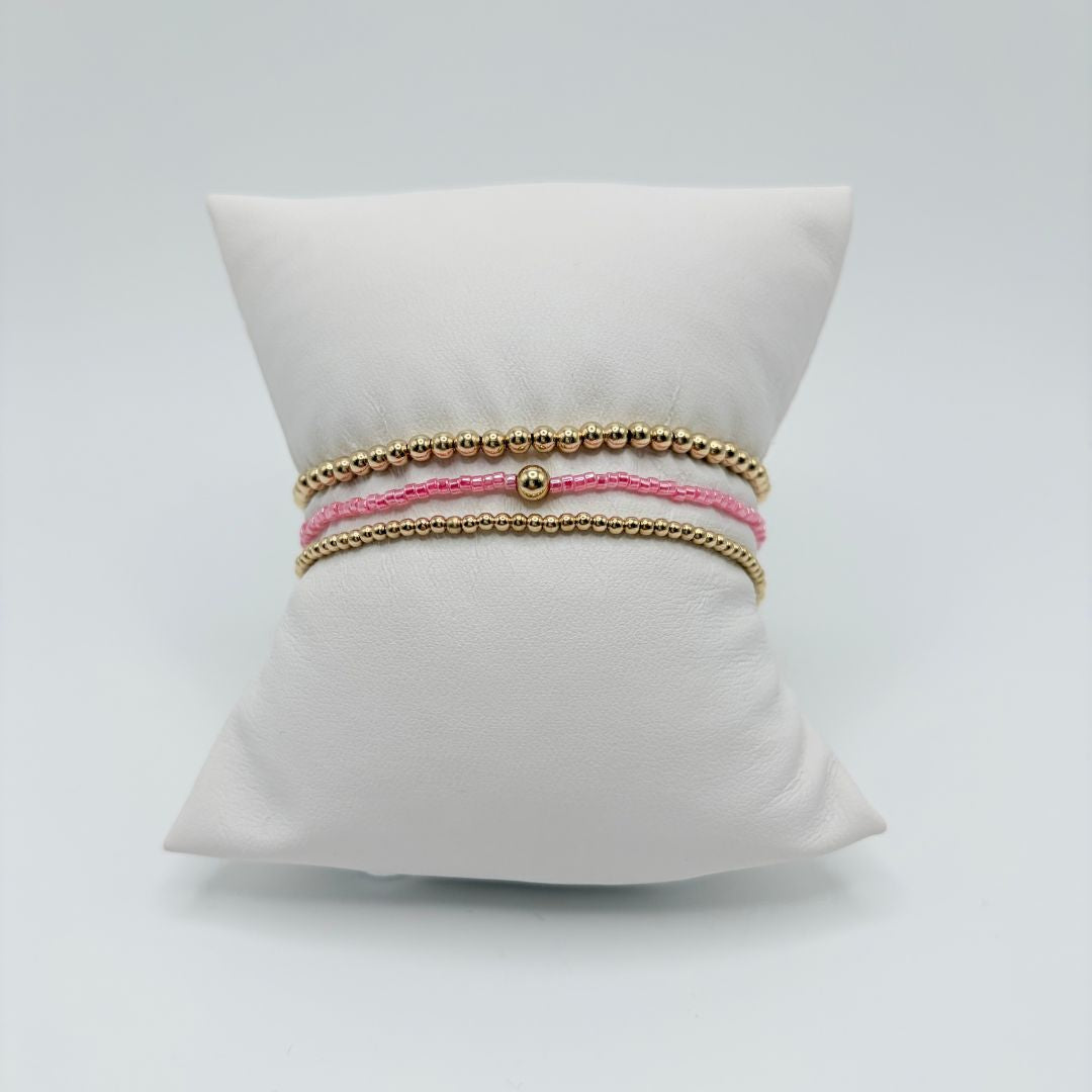 Three stacked bracelets on a white display pillow: pink seed bead bracelet with a gold bead between two gold beaded bracelets