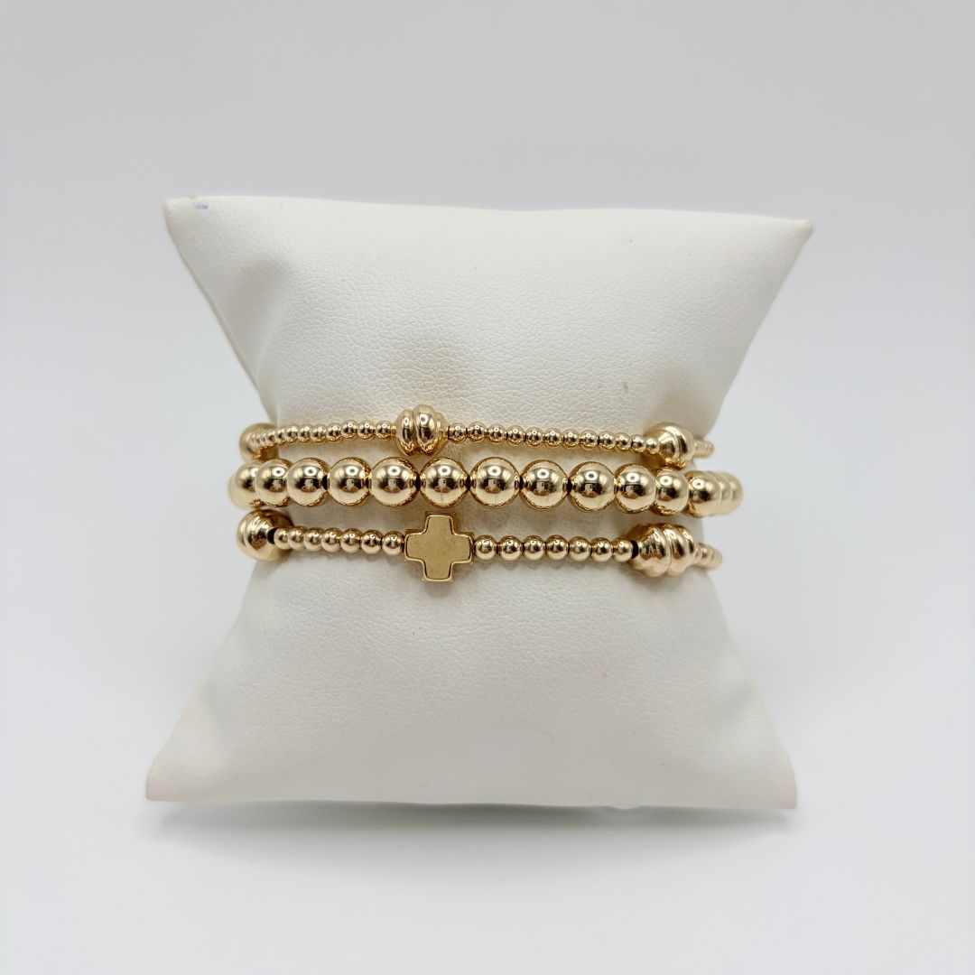 Three gold bracelets on a white pillow: large gold bead bracelet, gold cross bracelet and small gold beads with ribbed accents