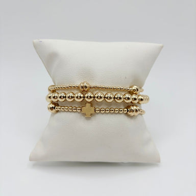 Three gold bracelets on a white pillow: large gold bead bracelet, gold cross bracelet and small gold beads with ribbed accents