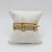 Three gold bracelets on a white pillow: large gold bead bracelet, gold cross bracelet and small gold beads with ribbed accents