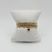 Four gold beaded bracelets in mixed bead sizes stacked on a white pillow, with a puffy gold heart charm