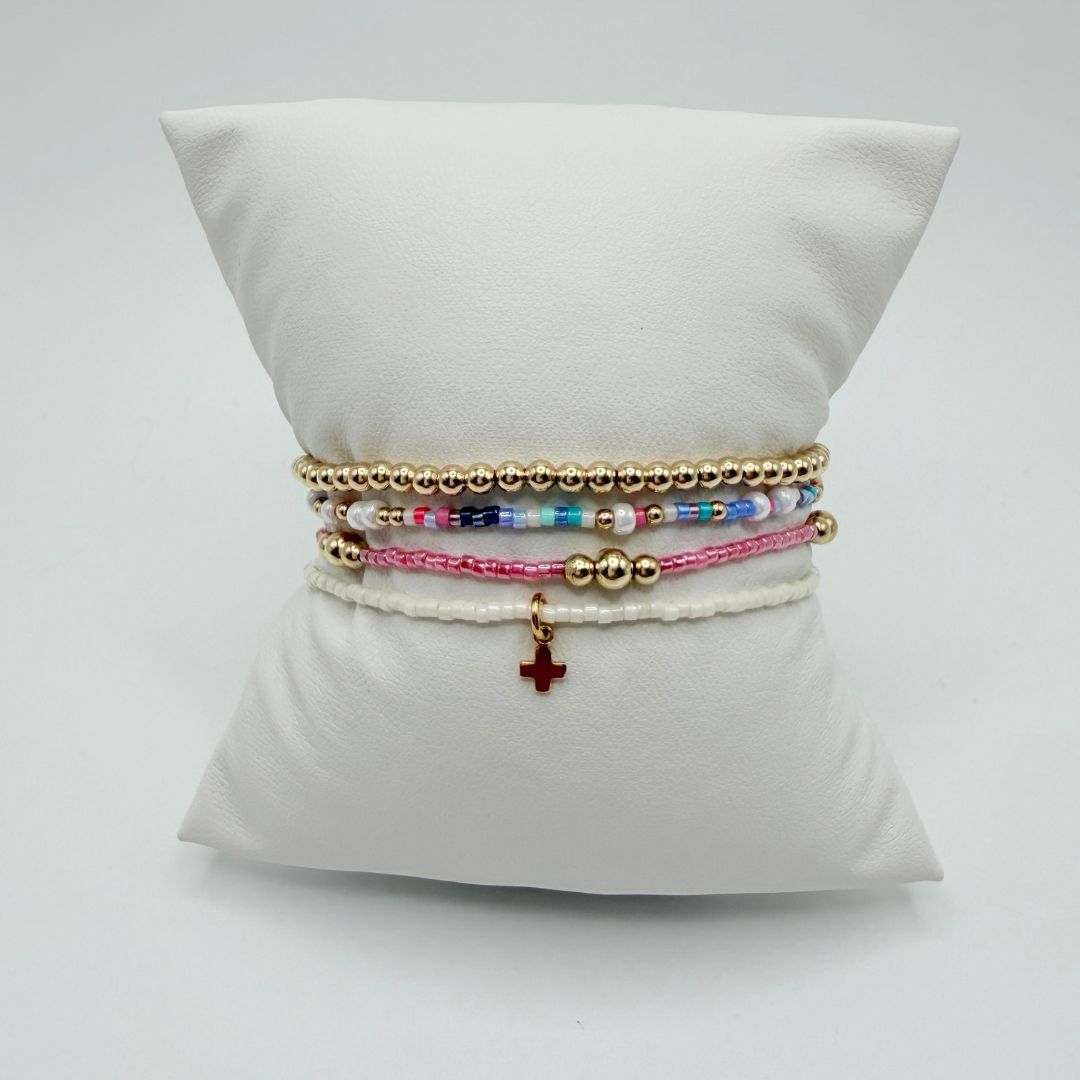 Four bracelets on a white pillow: gold beads, multicolor seed beads, pink seed beads and white seed beads with a red cross charm