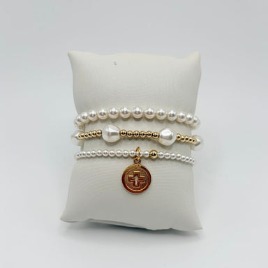 Three pearl and gold bracelets on a white pillow with baroque pearls and a round gold cross disc charm
