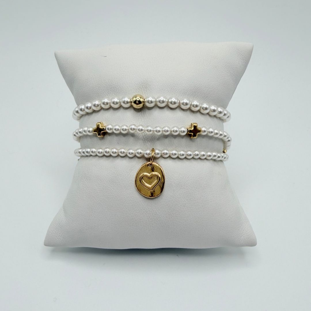 Three pearl bracelets on a white pillow with a gold bead, two gold crosses and a gold heart charm