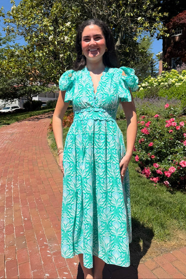 Woman in a teal dress standing outdoors with flowers and a path in the background