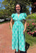 Woman in a teal dress standing outdoors with flowers and a path in the background