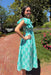 Woman in a green dress standing outdoors with trees and flowers in the background