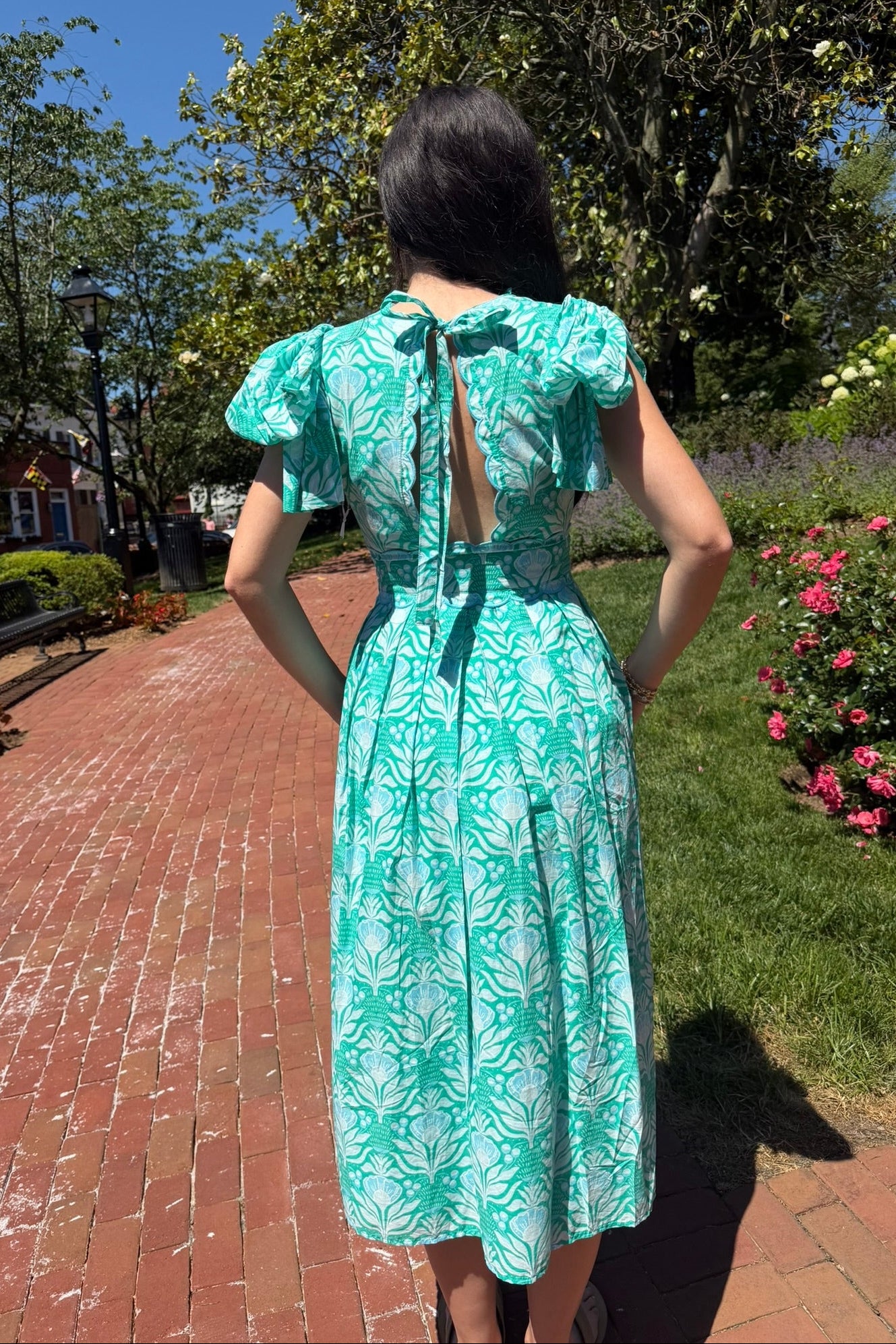 Woman in a teal dress with white patterns standing on a brick path with greenery and flowers in the background.