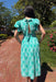 Woman in a teal dress with white patterns standing on a brick path with greenery and flowers in the background.