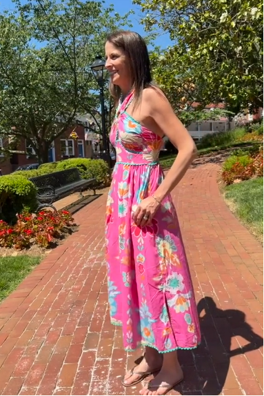 Woman in a pink floral dress standing on a brick path with trees and buildings in the background
