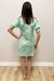 Woman wearing a light green floral dress against a plain wall.